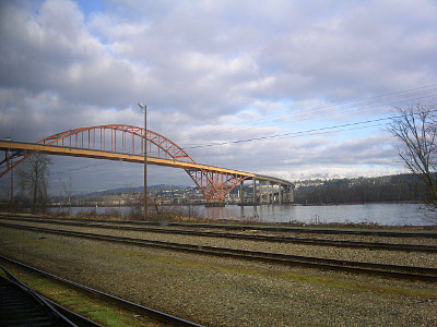 [The bridge is a yellow arch with the arch extending through the bridge deck to the water as part of the support. There are multiple empty train tracks in the foreground beside the river over which the bridge passes.]